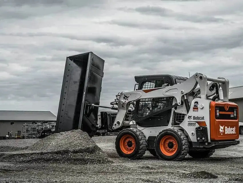 The image shows a Bobcat skid steer loader with a bucket attached to the front. The loader is parked on a dirt surface, and there is a pile of dirt in front of the bucket.
