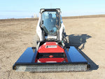 Frontal view of a skid steer equipped with a red and black brush mower attachment, positioned on a plowed field, with the operator visible in the cabin, ready to begin land clearing work.