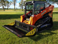 Thumbnail for  A Kubota SVL90 skid steer loader with an Rock bucket attachment parked on a grassy field. The skid steer loader has its front bucket raised and is turned slightly to the right. The grass is green and there are a few trees in the background.