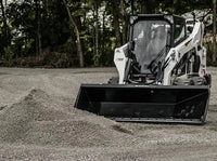 Thumbnail for The image shows a skid steer with a large bucket on top of a pile of gravel. The skid steer is white and black, and the bucket is black. The pile of gravel is in a vacant lot, and there is a row of trees in the background.