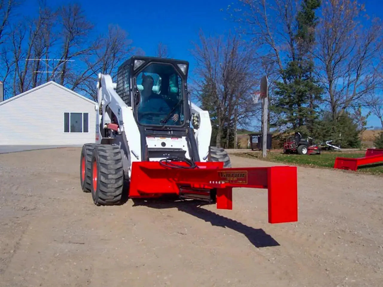 A TM Manufacturing Pro 30 skid steer log splitter attachment in the snow. The splitter is bright red and has a black metal frame with red hydraulic hoses and a control panel. The text “T.M. SKID SPLITTER 30″ Pro” is written in white on the side of the splitter.