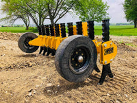 Thumbnail for A yellow tracked skid steer loader with black rubber tracks and a yellow plow attachment parked on a dirt construction site. The plow attachment has six curved teeth and tapers to a point at the front. Behind the skid steer loader is a pile of dirt and a blue dumpster. 