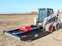 Thumbnail for A skid steer loader equipped with a brush mower attachment working on a barren field, highlighting the mower's broad cutting deck and the skid steer's muddy wheels, against a backdrop of a clear sky and distant farm structures.
