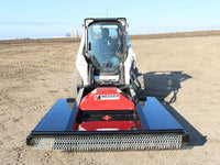 Thumbnail for Frontal view of a skid steer equipped with a red and black brush mower attachment, positioned on a plowed field, with the operator visible in the cabin, ready to begin land clearing work.