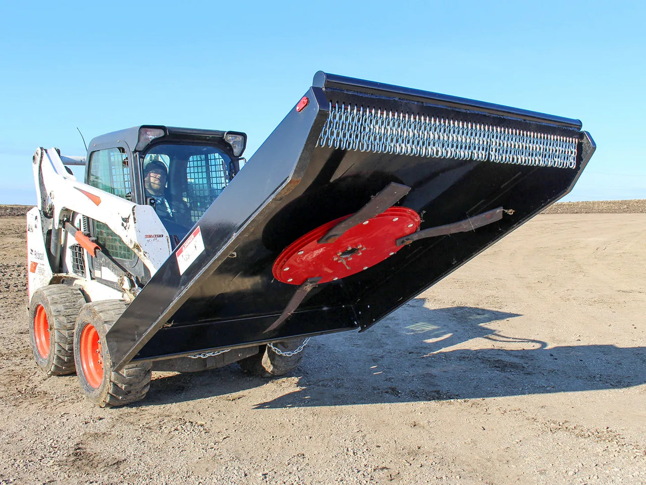 A skid steer equipped with a 72" brush cutter attachment from Messer Attachments. The brush cutter, held aloft, reveals its robust red cutting blades and protective chain guard, showcasing its heavy-duty design for effective land clearing and brush cutting. The skid steer operator is visible inside the cab, demonstrating the equipment's operational setup.